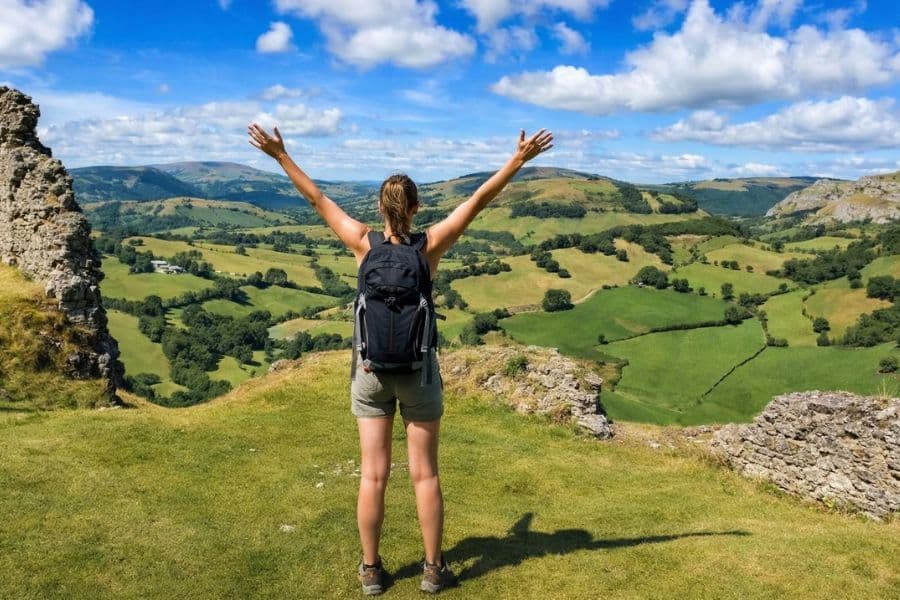 Hiker with a backpack stands on a grassy hilltop with arms raised, looking out over rolling green valleys and patchwork fields under a bright blue sky. Scenic viewpoint that fits a roundup of hiking trails in Europe.