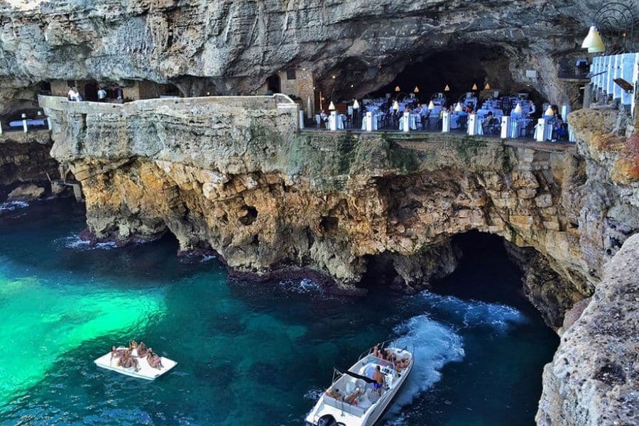 Seaside cave restaurant built into dramatic cliffs above turquoise water with boats below showcasing unique villages in Italy.