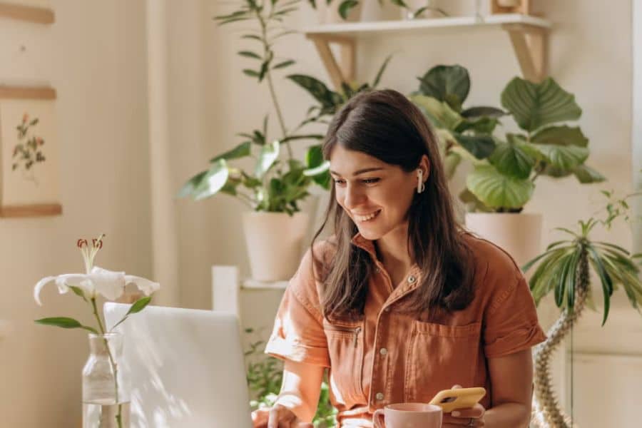Woman smiling at a laptop in a bright plant filled home office while holding her phone. This scene fits work from home jobs content by showing a relaxed remote work setup with natural light and a comfortable workspace.