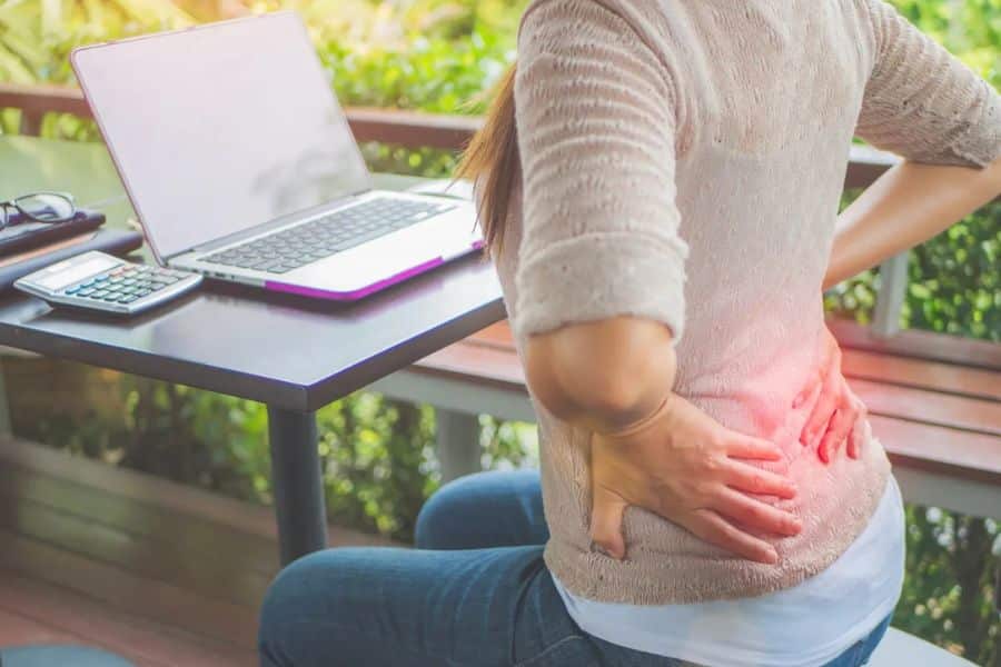 Woman seated at an outdoor desk with one hand pressed against her lower back while working on a laptop. The photo shows back pain linked to sitting position during desk work.