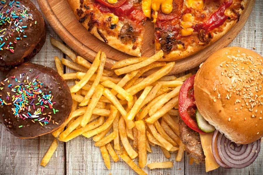 Top view of a fast food spread with chocolate frosted donuts covered in rainbow sprinkles. Fries are piled between a cheeseburger and sliced pizza to show a mix of highly processed foods that may come up in conversations about hunger hormones.