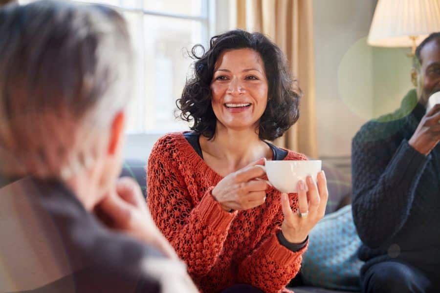 Woman in a rust colored sweater smiles while holding a white mug and talking with two other people in a bright living room. This could support a micro habit around taking a quiet tea break or connecting with friends.