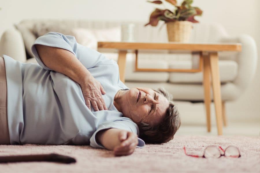 Older woman lies on a living room floor clutching her chest with her eyes shut in pain while her glasses rest nearby on the carpet. This scene shows a sudden medical emergency at home that could be confused with stroke symptoms.