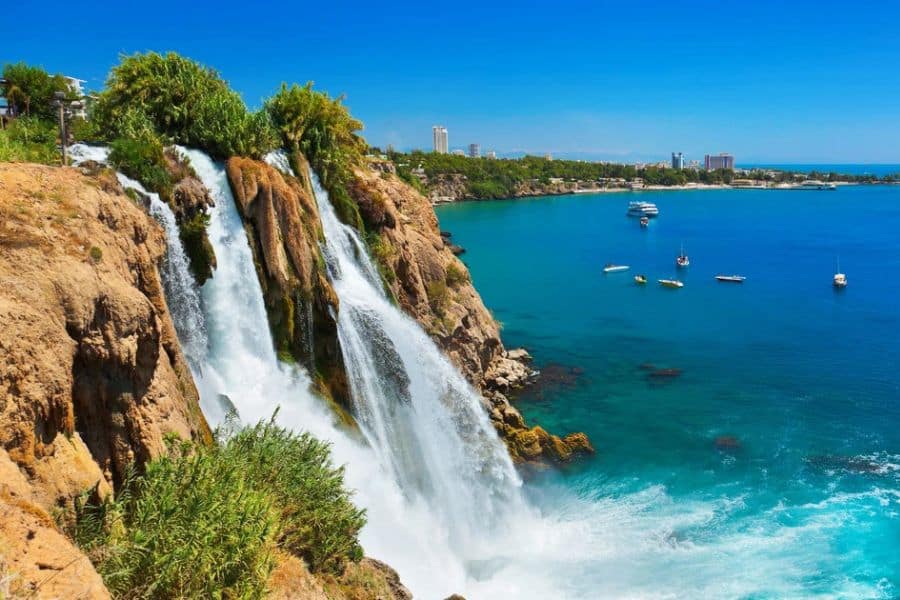 A wide waterfall pours over a rocky cliff covered in greenery into bright turquoise sea while small boats float in the bay nearby. This highlights vacation destinations with a dramatic mix of coastline, clear water, and a scenic natural landmark.