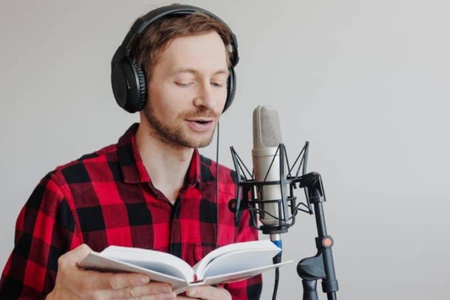 Man in headphones reads from an open book into a studio microphone against a plain background. This supports work from home jobs content related to voiceover work or remote audio recording.