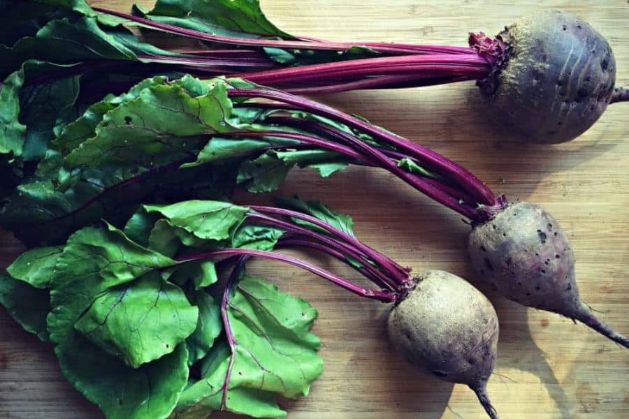 Three beets with dusty roots and long red stems lie on a wooden board with their leafy tops attached. The photo highlights beet greens as edible leaves as well as the root vegetable itself.