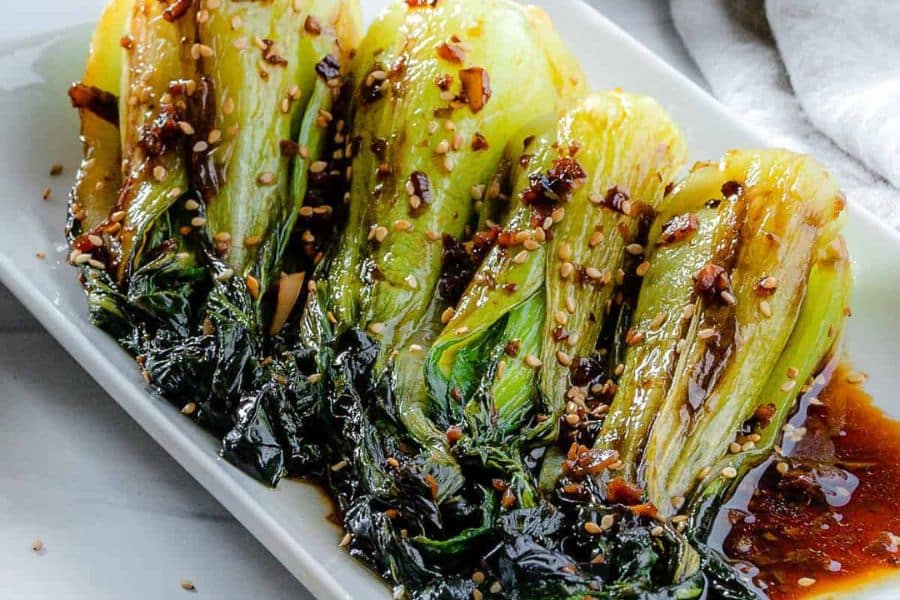Cooked bok choy is arranged in a row on a white plate and coated in a glossy dark sauce with sesame seeds and chili bits. The close crop emphasizes the tender green leaves and pale stems.