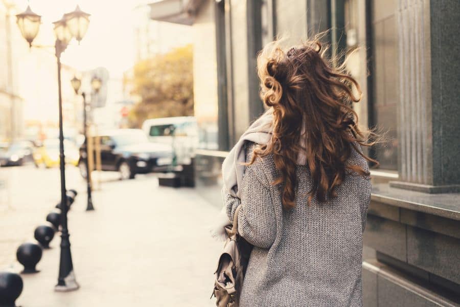 Woman with long wavy hair walks down a sunlit city sidewalk wearing a gray sweater and scarf. The photo suggests a simple daily walk as part of a healthy routine.