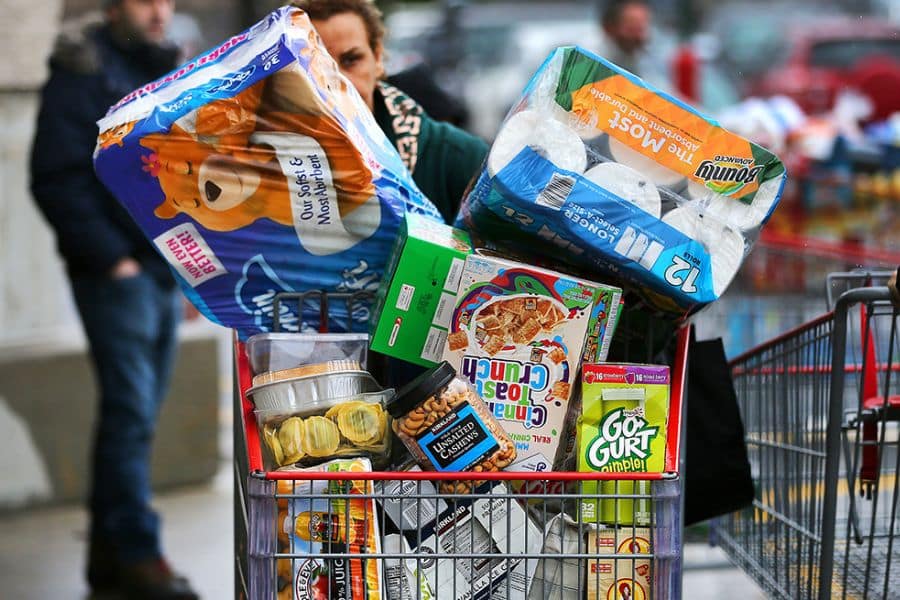 Grocery cart piled high with household supplies and packaged foods including paper towels cereal yogurt and cashews as a shopper pushes it through a store. This fits content about how to save money on groceries by showing a large stock up shopping trip.