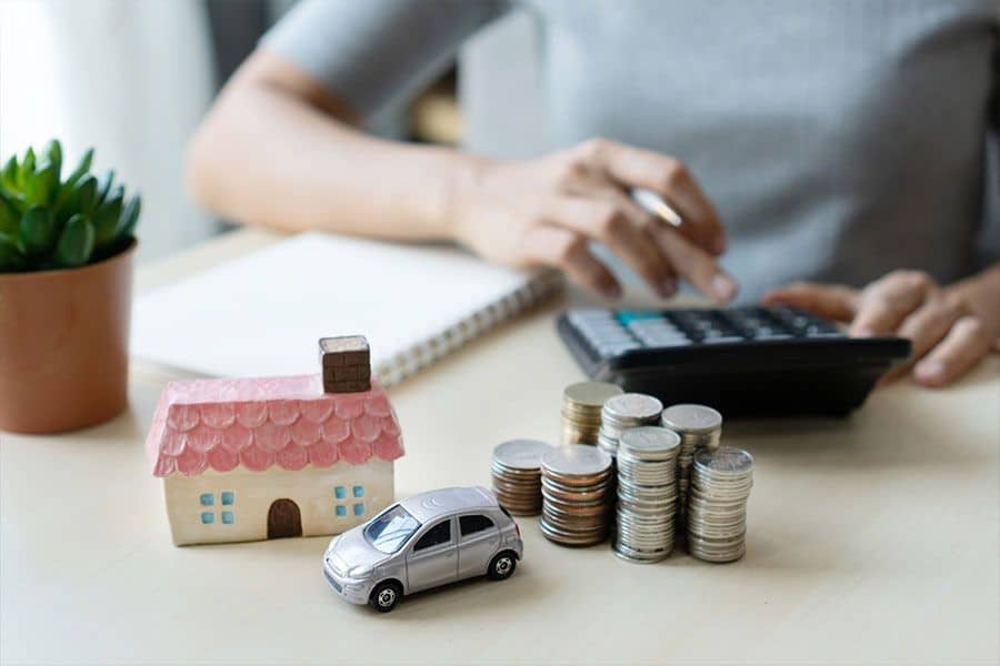 Small model house and silver toy car sit beside stacks of coins on a desk while a person uses a calculator in the background. This setup represents household and car related expenses and fits a topic about how to save money on insurance.