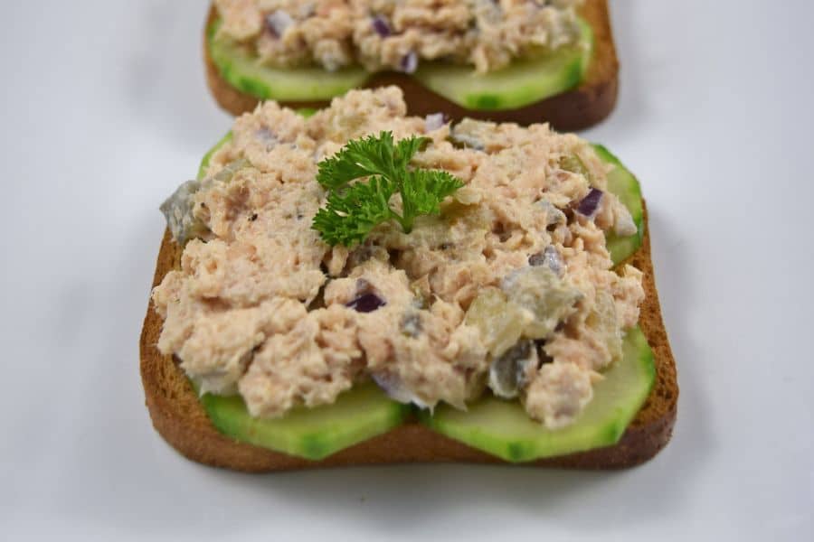 Two open faced sandwiches are topped with cucumber slices and a chunky tuna salad mixture with diced red onion and a parsley garnish. The front toast is in sharp focus against a plain white background.