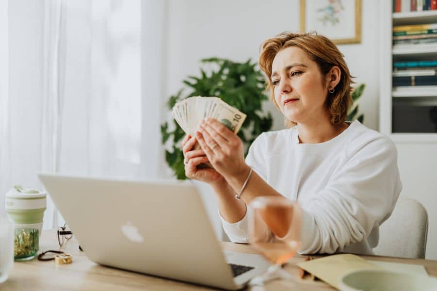Woman sitting at a desk with an open laptop while counting a fan of cash in her hands. The scene suggests closely tracking income and expenses at home.