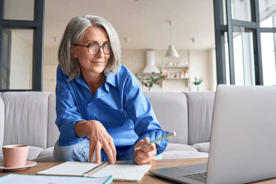 Gray haired woman in a blue shirt sits on a sofa with a notebook and pen while looking at a laptop. The scene suggests a ChatGPT assisted video call, online learning session, or remote work task in a bright home setting.