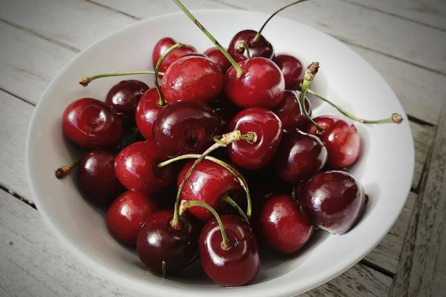 White bowl filled with fresh dark red cherries on a light wooden table. The close up highlights cherries as one of the foods you should eat for a simple fruit based snack.