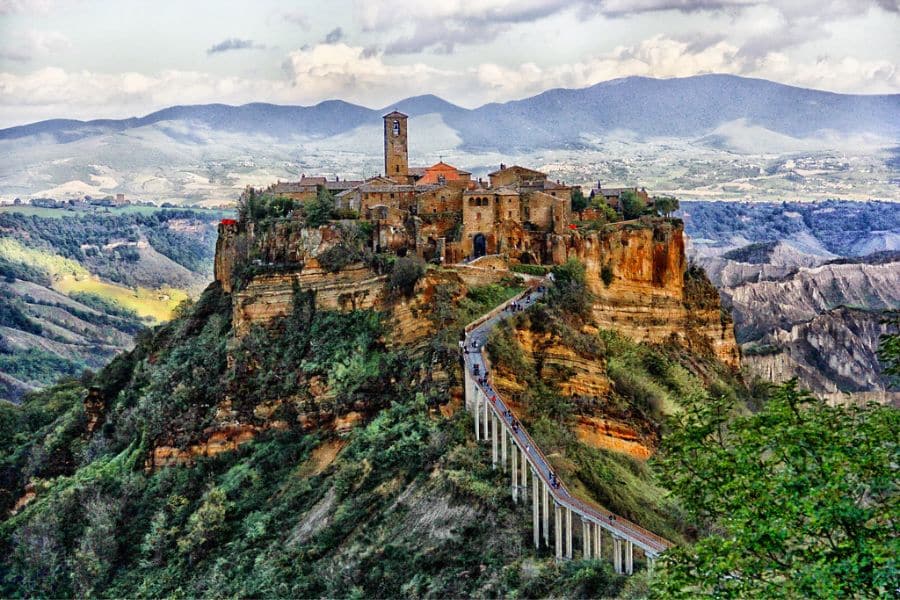 Historic hilltop village perched on a rocky cliff with a long pedestrian bridge leading to stone buildings representing medieval villages in Italy.