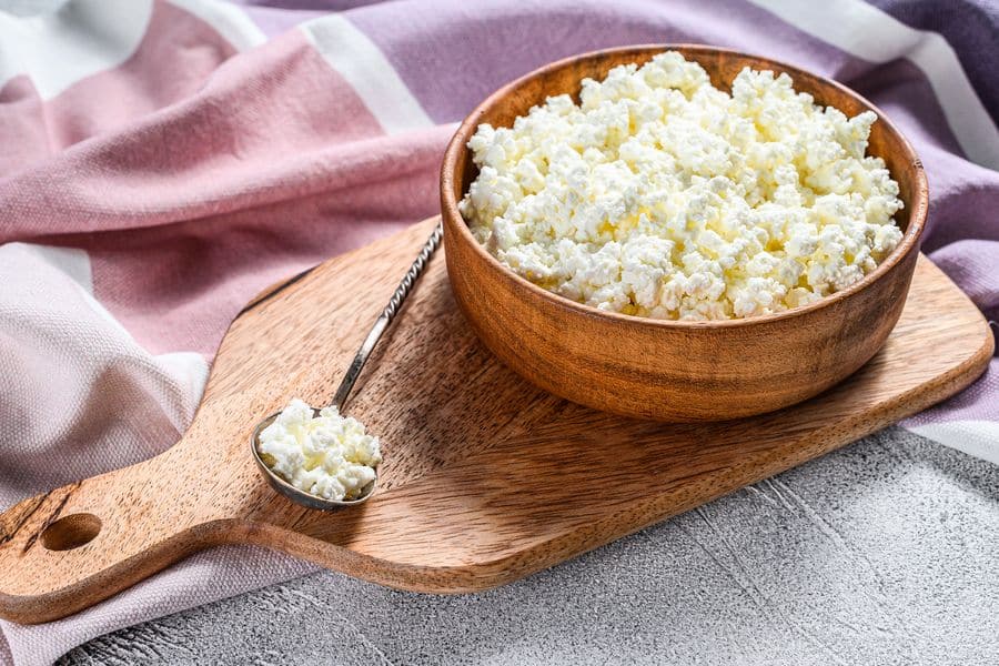 Wooden bowl filled with cottage cheese sits on a cutting board with a spoonful beside it and a pink striped cloth draped in the background. The texture of the curds is clearly visible in this simple tabletop shot.