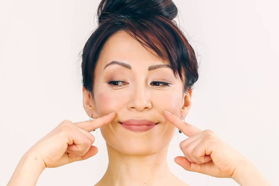 Woman with her hair in a bun smiles slightly while pressing both index fingers into the apples of her cheeks against a plain light background. The pose demonstrates a facial yoga cheek exercise focused on lifting and toning the face.