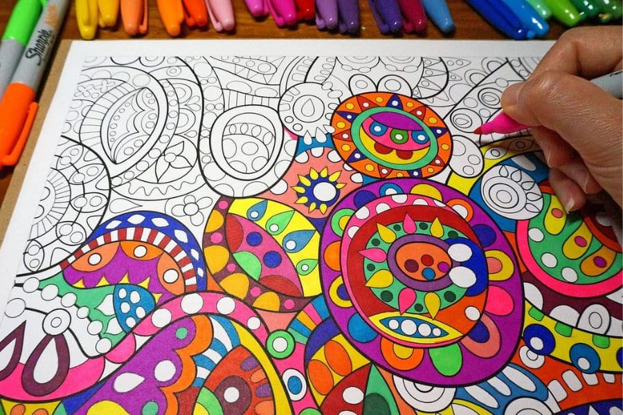 A hand colors an intricate abstract coloring page with bright markers while uncapped pens line the top edge of the table. This could represent using AI to make money by creating printable coloring pages or other digital art products.