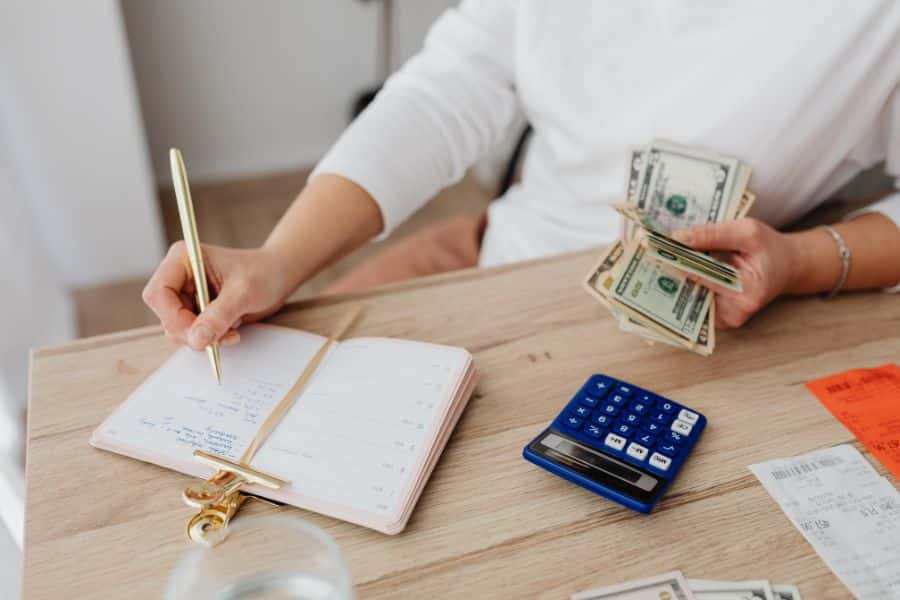 Person writing in a planner while holding cash beside a blue calculator, receipts, and papers on a wooden table. This captures hands on budgeting and planning around limited money.