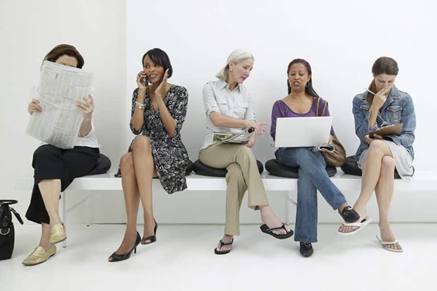 Five women sit side by side in a waiting room while reading a newspaper talking on the phone using a laptop and writing on a notepad. Their different sitting positions suggest a range of everyday seated postures.