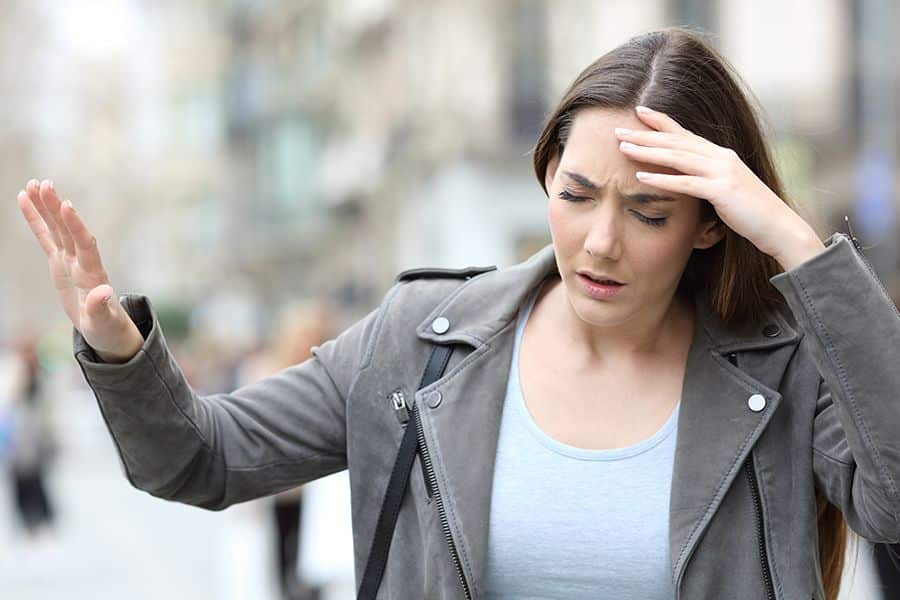 Young woman standing outdoors looks dizzy and unsteady with one hand pressed to her forehead and the other raised for balance. Her expression and posture suggest disorientation that may be associated with stroke symptoms.