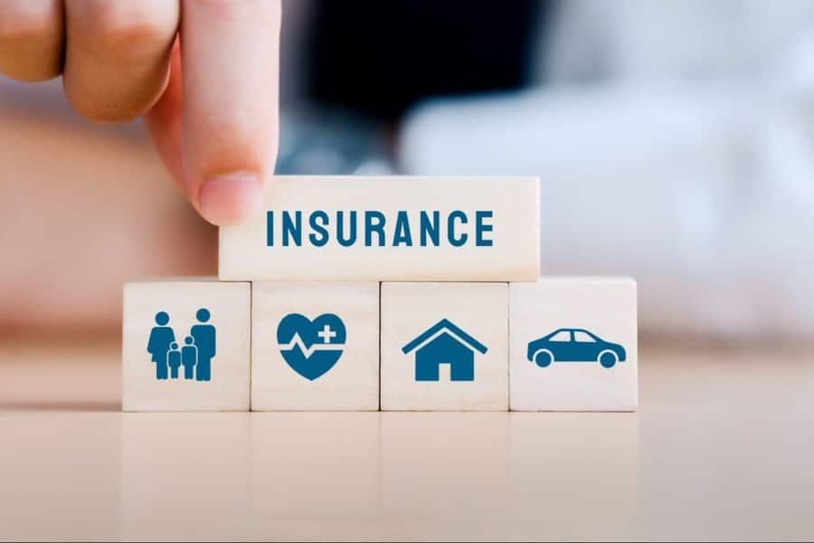 A hand places a block labeled "INSURANCE" on top of four wooden blocks showing family health home and car icons. The arrangement highlights different types of coverage grouped together in one simple visual.