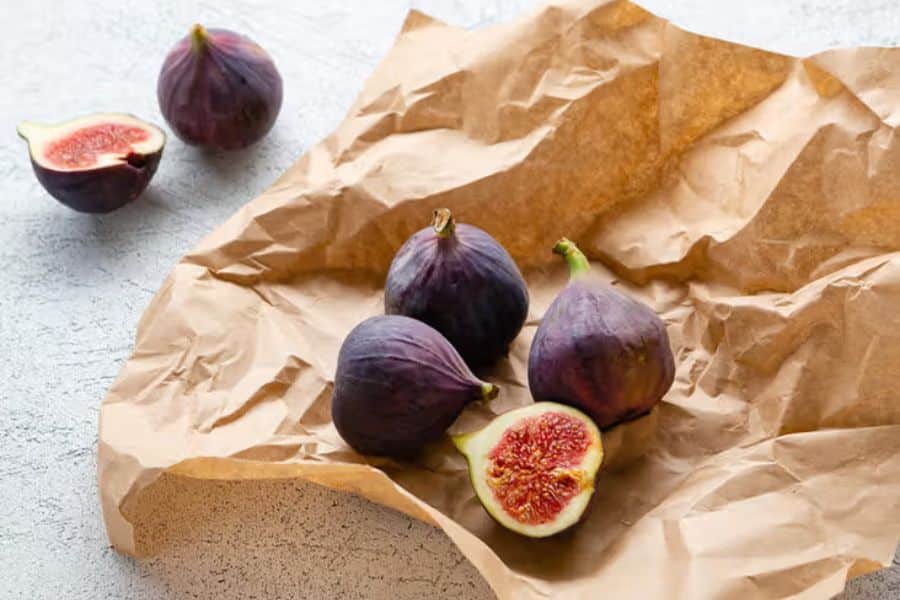 Several fresh purple figs rest on crinkled brown paper with two cut open to show their deep pink centers. The neutral background keeps the focus on the fruit and its rich color.