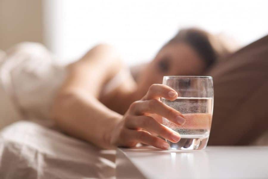 Person lying in bed reaches toward a glass of water on a bedside table. This works well for a micro habit about drinking water first thing in the morning.