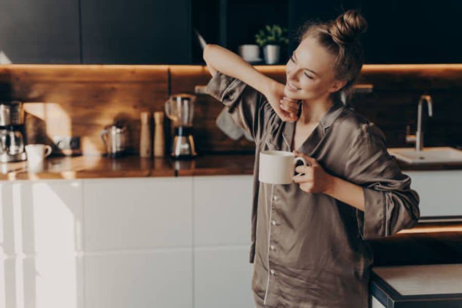 A woman in satin pajamas leans against a kitchen counter while holding a mug in warm morning light. This quiet coffee moment fits a post about morning rituals destroying your metabolism.