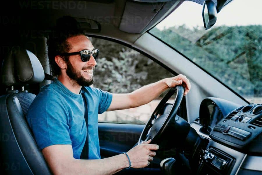 Man wearing sunglasses smiles while driving a van with both hands on the steering wheel. His upright sitting position shows posture while seated in a vehicle.