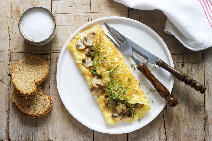 Mushroom omelet folded on a white plate with sliced bread and a small cup on a rustic wooden table. The meal suggests a quick nourishing breakfast choice.