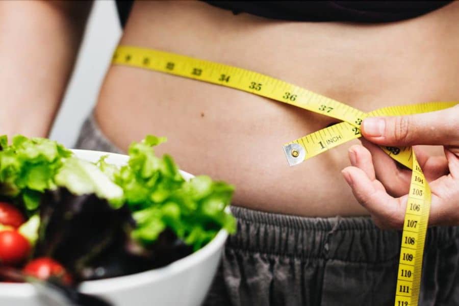 Close up of a person measuring their waist with a yellow tape measure while a bowl of salad sits blurred in the foreground. The photo represents dieting and body measurement during a losing weight journey.