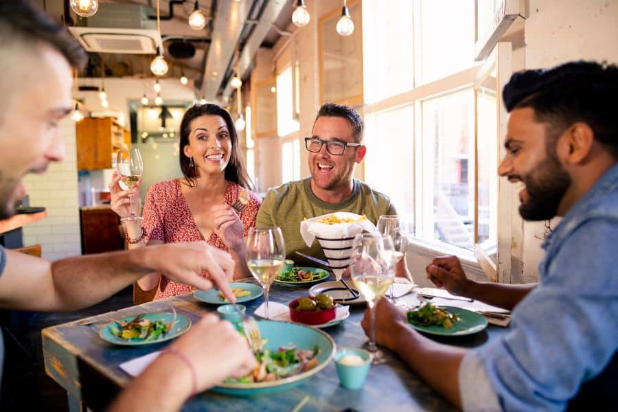 Four friends sit around a restaurant table laughing over drinks plates of food and a basket of fries near a bright window. This supports content about how to save money while dining out or socializing on a budget.