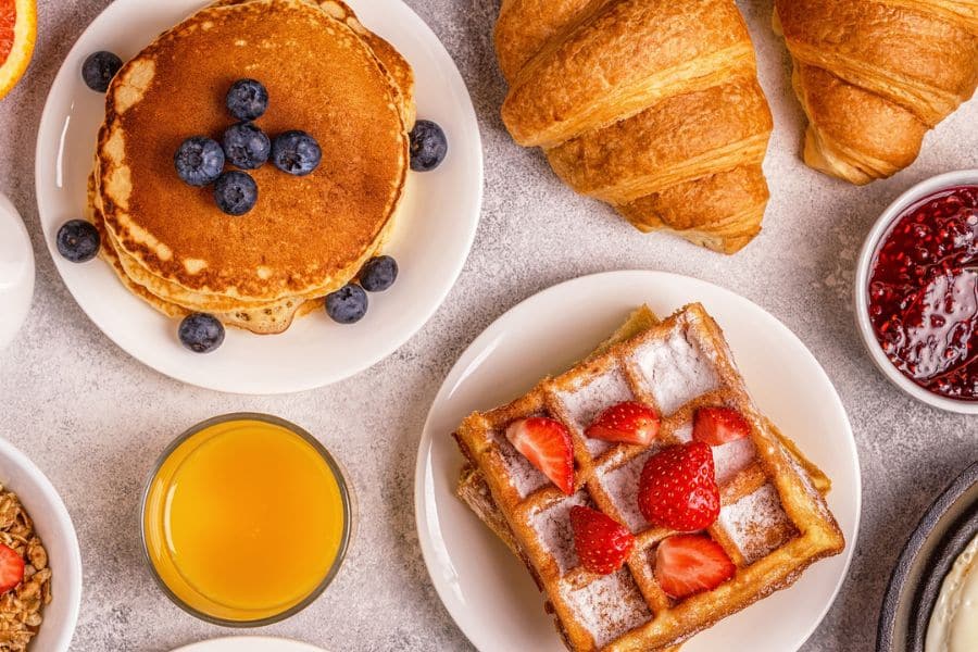 Overhead view of a breakfast spread with blueberry pancakes, waffles topped with strawberries, croissants, orange juice, and jam. The rich mix of pastries and sweet breakfast foods highlights a heavy morning meal.