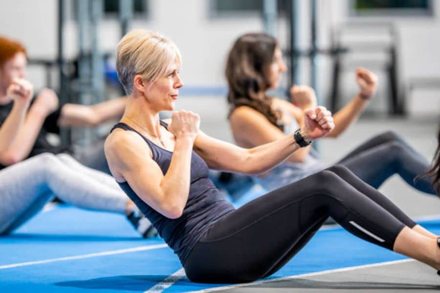 A group fitness class does seated core exercises on blue mats in a gym. A silver haired woman in the foreground holds her fists up while leaning back in a controlled movement.