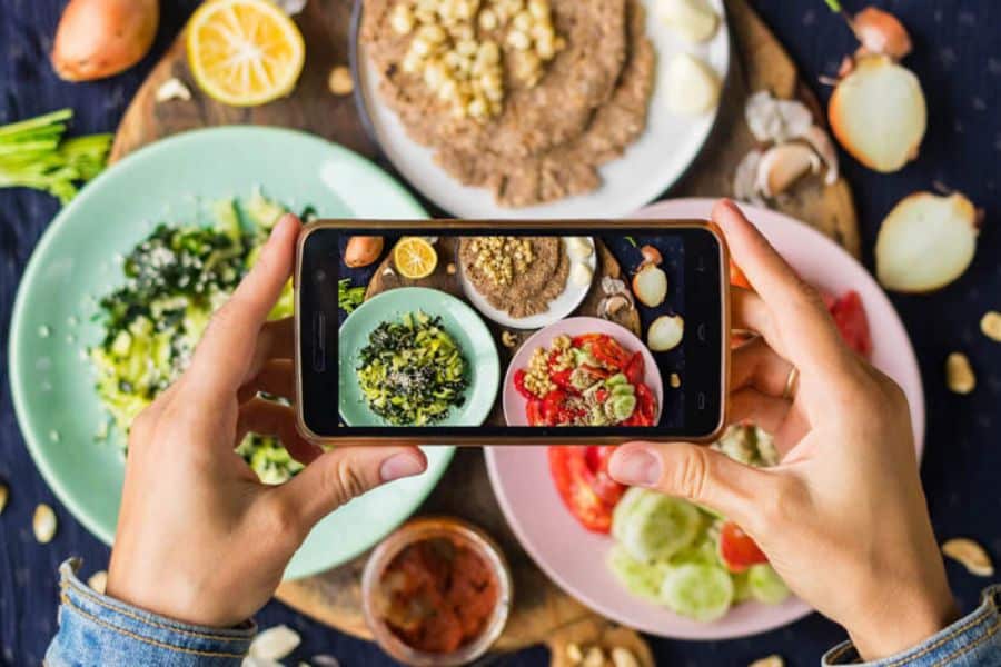 Hands hold a phone above colorful plates of food while taking a photo from overhead. This fits work from home jobs content about food blogging social media management or content creation from home.