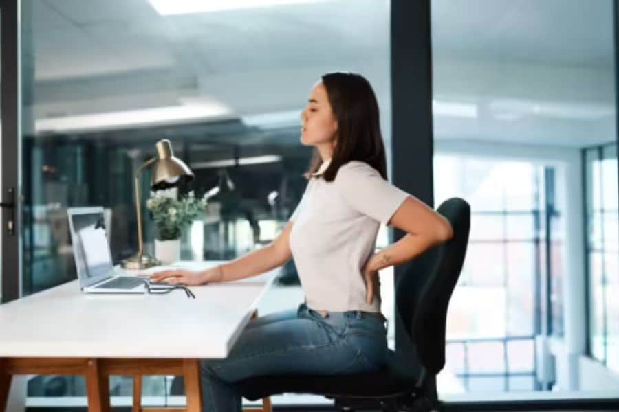 Woman sits at an office desk with one hand on her lower back and the other near a laptop. The scene highlights discomfort caused by sitting position at work.