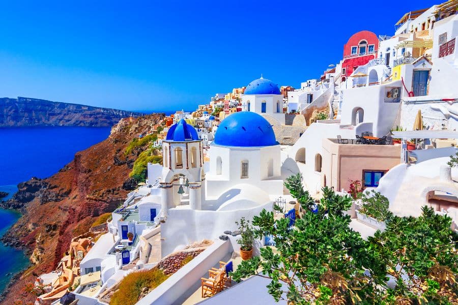 Whitewashed buildings with rounded walls and bright blue domes cluster along a steep seaside cliff above deep blue water. This sunlit Santorini view reflects the relaxed coastal beauty often associated with retiree hotspots in Europe.