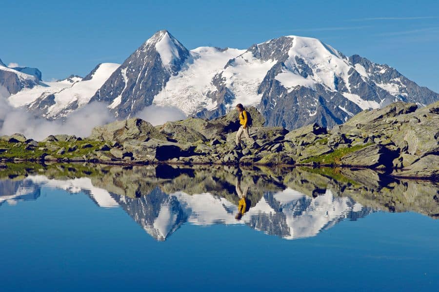 Hiker walking beside a crystal clear alpine lake with snow capped mountains in the distance within European National Parks.