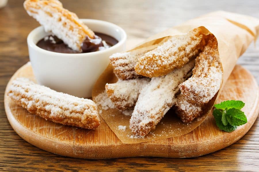 Plate of churros dusted heavily with powdered sugar beside a cup of chocolate dipping sauce and a sprig of mint. The sweet fried pastry is presented as a rich dessert or snack.