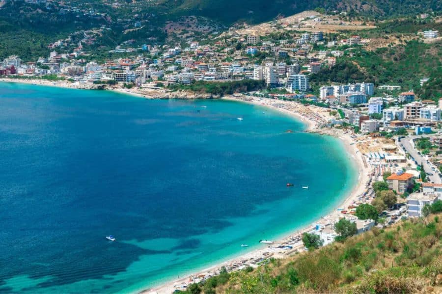 A curved beach with bright blue water wraps around a coastal town built along green hillsides. This fits vacation destinations content by showing a sunny bay, swimmable shoreline, and a dense seaside resort area.