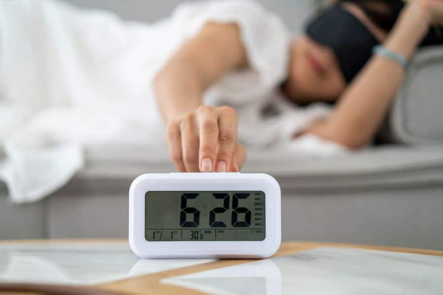 A person wearing a sleep mask reaches toward a digital bedside clock that reads "6:26" while lying in bed. The scene shows an early wake up moment in a soft blurred bedroom.