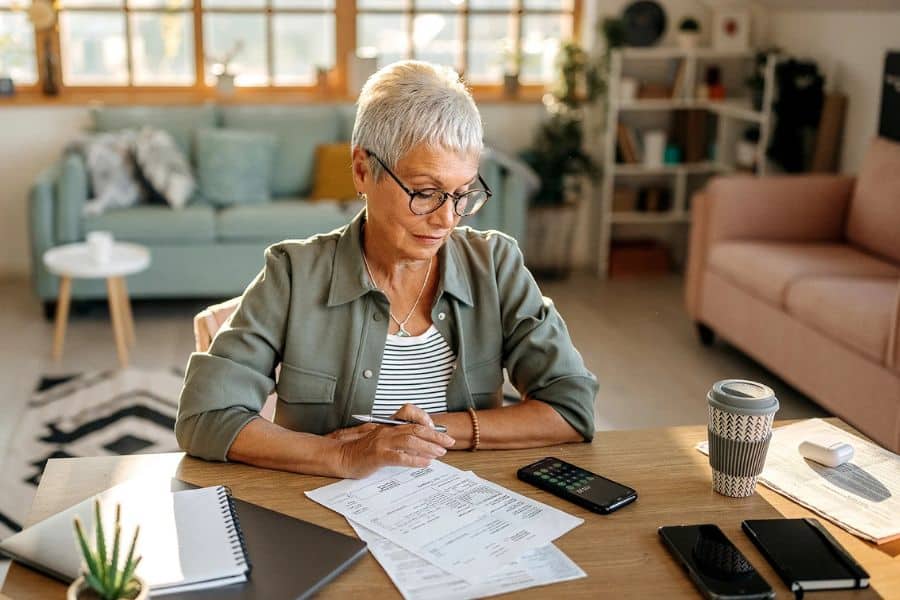 A mature woman with glasses sitting at a desk in her living room looking at paperwork