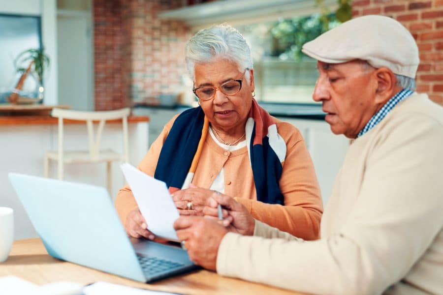 Older couple reviewing paperwork together at a kitchen table with a laptop open in front of them. This retirement planning scene shows them focused on documents and discussing finances at home.