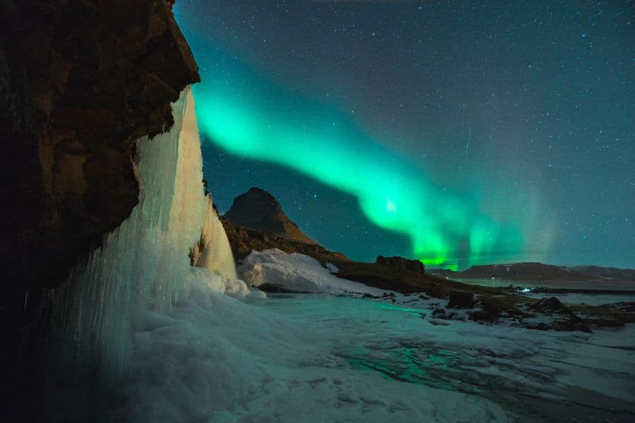 Green northern lights sweep across a dark star filled sky above an icy shoreline and a frozen waterfall at the left edge of the scene. Snow covered ground and shadowy mountains make the landscape feel remote and dramatic.