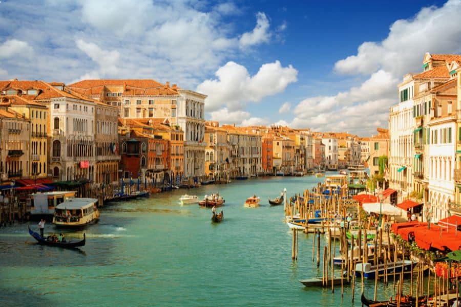 A broad canal runs between historic buildings with warm orange roofs while gondolas and small boats drift across the water. Wooden mooring poles and waterfront facades create a classic Venice cityscape.