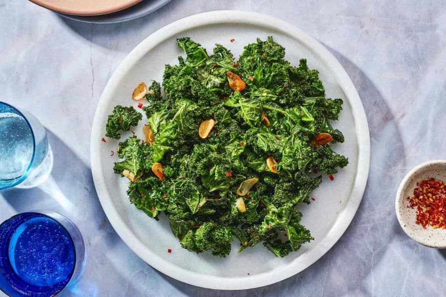 Plate of sautéed kale is topped with sliced garlic and red pepper flakes on a light stone surface. A blue drinking glass and a small bowl of extra pepper flakes frame the plate from the side.