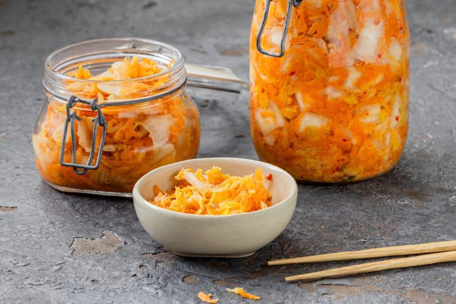 Two glass jars and a small bowl are filled with bright orange and white fermented vegetables on a gray surface. The photo highlights probiotic rich fermented food that may be included in a memory decline diet.