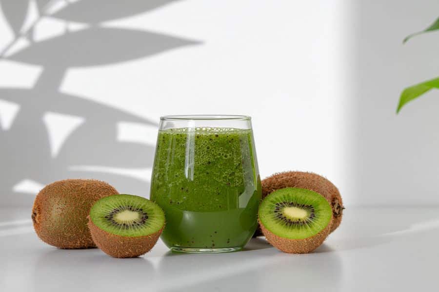 Glass of green kiwi smoothie surrounded by whole and halved kiwis on a white surface with soft plant shadows in the background. This photo shows a fresh kiwi drink as a bright nutrient rich choice.