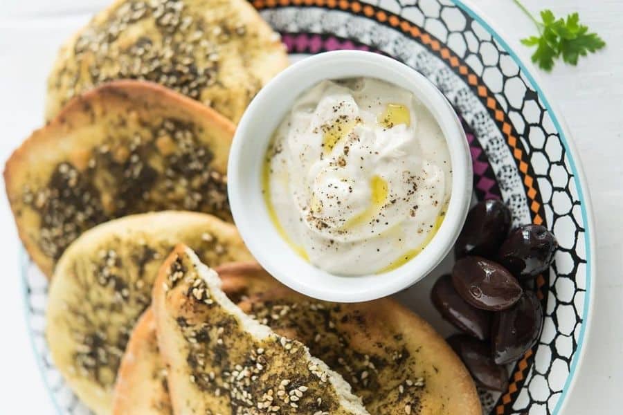 Plate of laneh topped flatbread served with a bowl of creamy white dip and a handful of dark olives. The overhead view highlights a simple spread of Mediterranean foods with contrasting textures and colors.